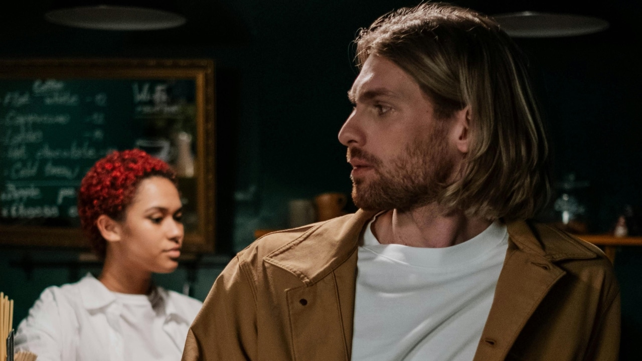 A man with shoulder-length hair looks sideways while a woman with red hair stands behind him.