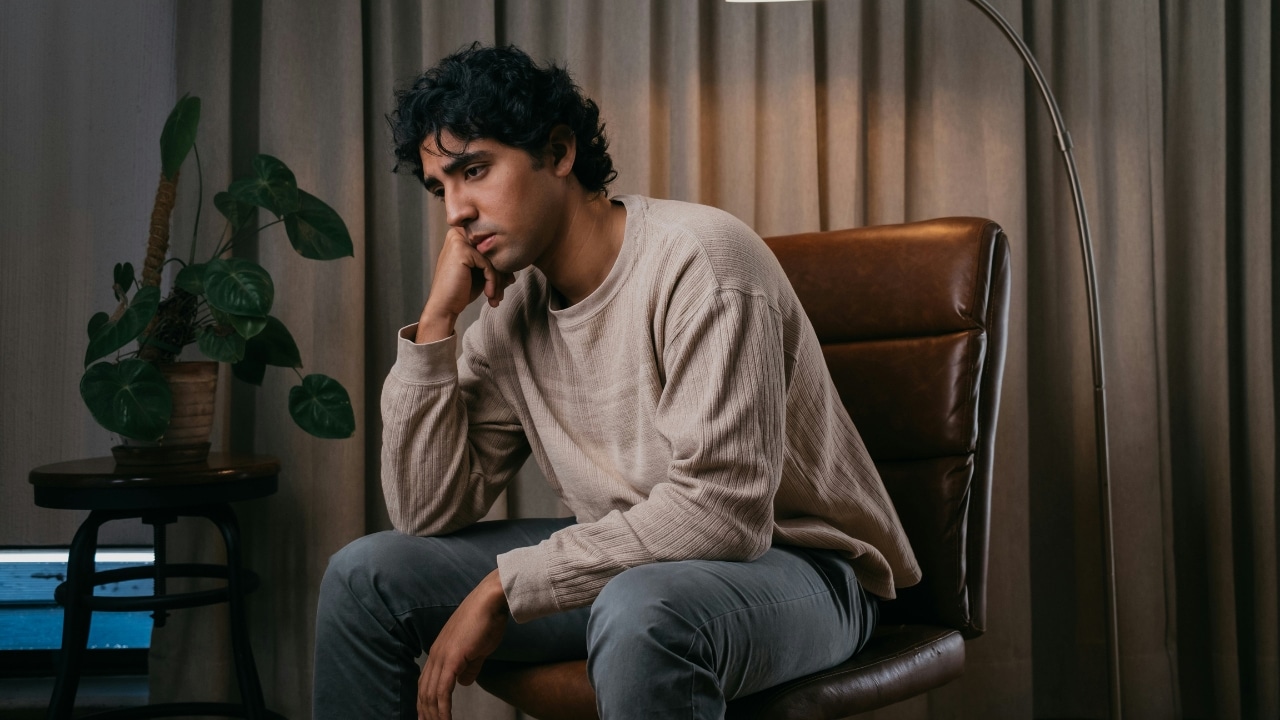 A man with curly hair sits on a leather chair, looking down with a somber expression.