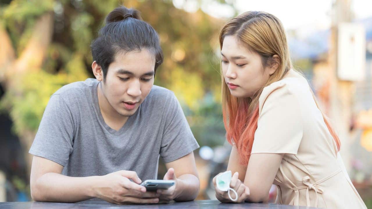 A man looks down at a smartphone while a woman watches him with a neutral expression.