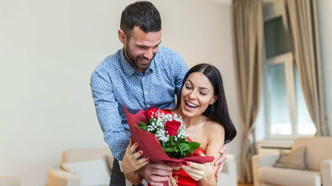 A smiling man giving a bouquet of red roses and baby's breath to surprise a woman.