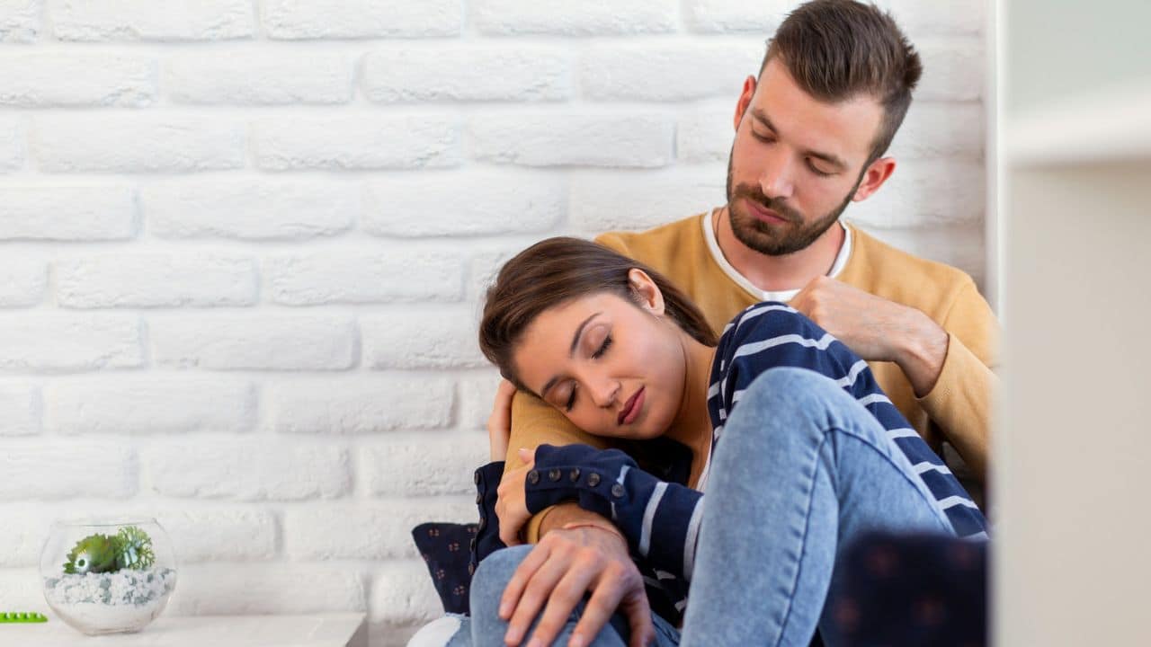 A woman rests her head on a man’s shoulder against a white brick wall background.