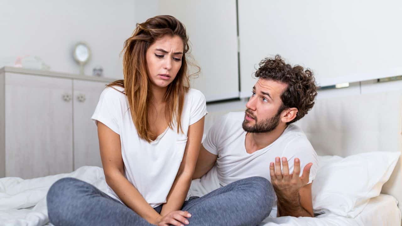 A woman sits on a bed looking down while a man gestures with one hand.