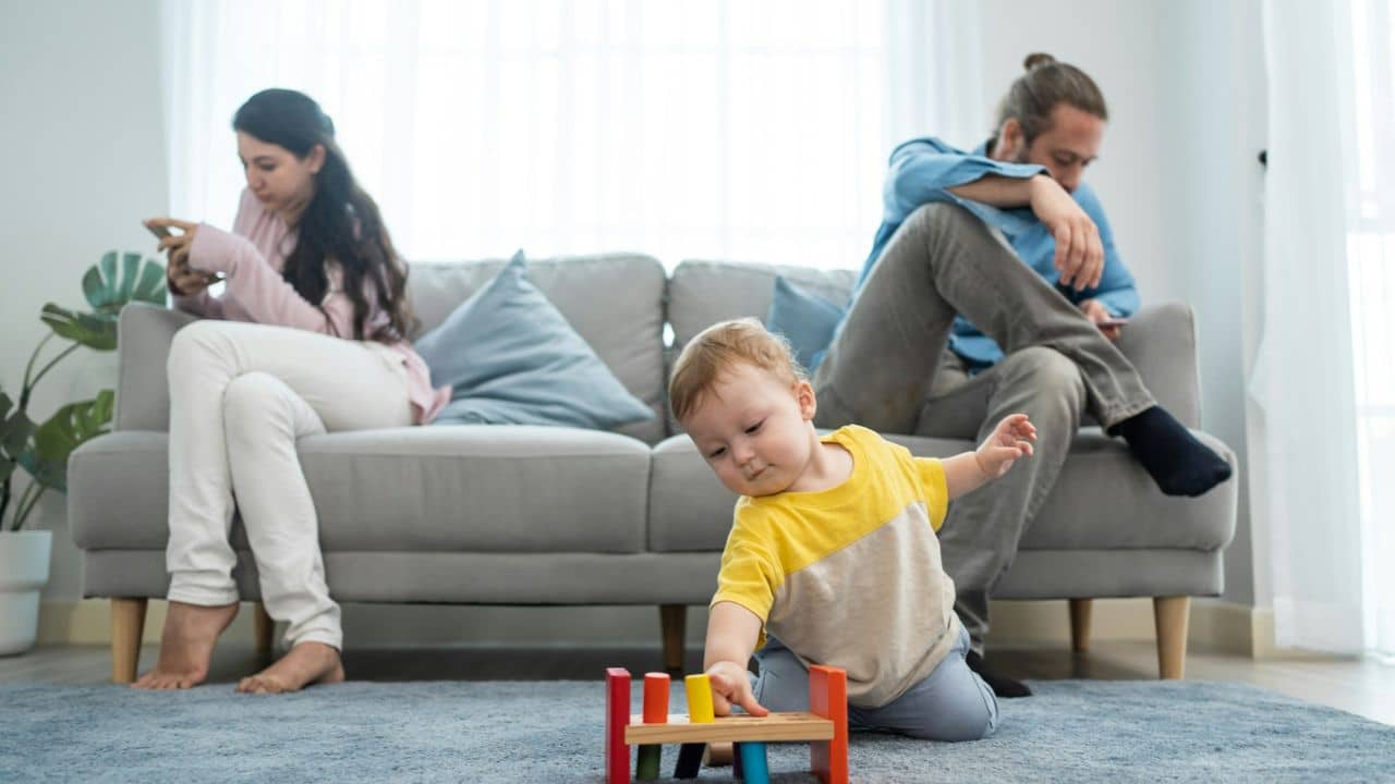 A child plays with blocks while two adults sit apart on a sofa using phones.