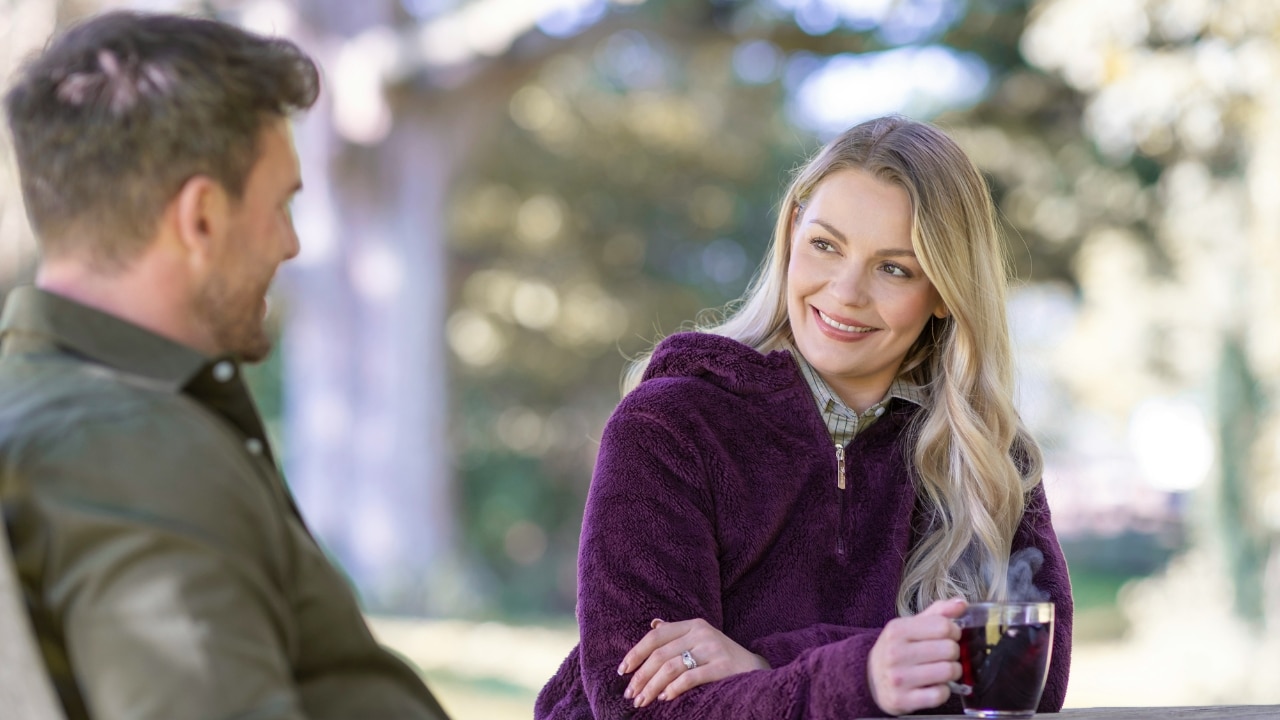 A smiling woman in a purple fleece jacket sits outdoors holding a mug near a man.