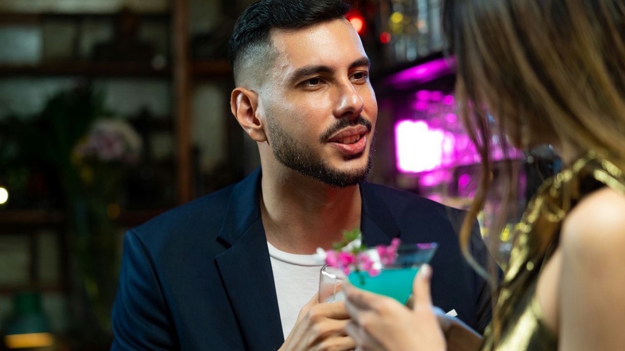 A man with a beard looks at a woman while holding a glass in a bar.