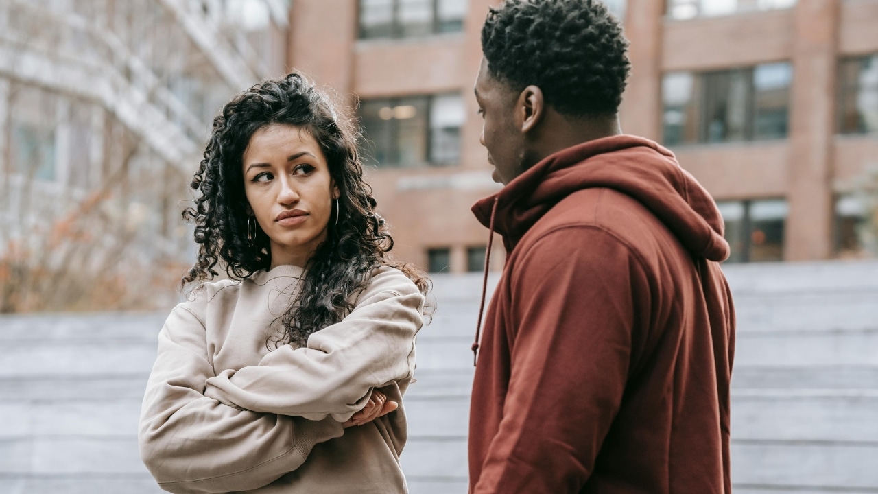 A woman with crossed arms looks sideways at a man in a red hoodie.