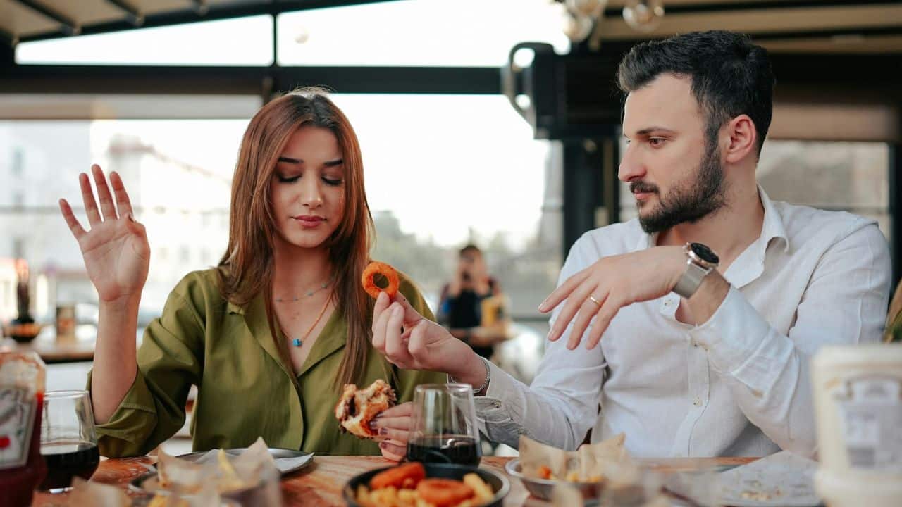 A man offers an onion ring to a woman while eating lunch at an outdoor table.