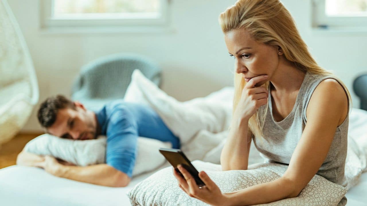 A woman sits on a bed looking at a smartphone while a man lies behind her.