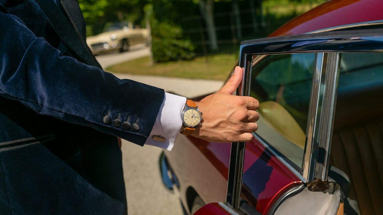 A man in a velvet suit opens a car door, showing a watch on his wrist.