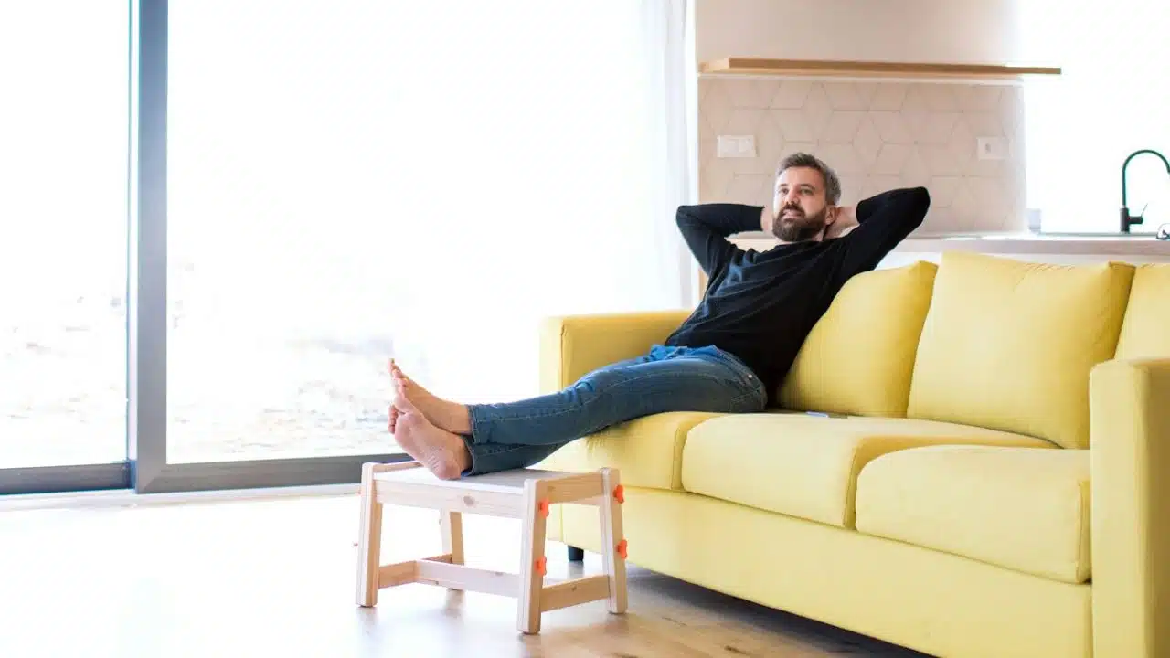 A man reclines on a yellow sofa with his hands behind his head and feet up.