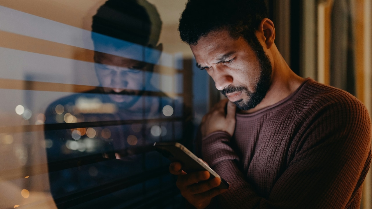 A man with a beard looks down at a glowing smartphone near a reflective window.