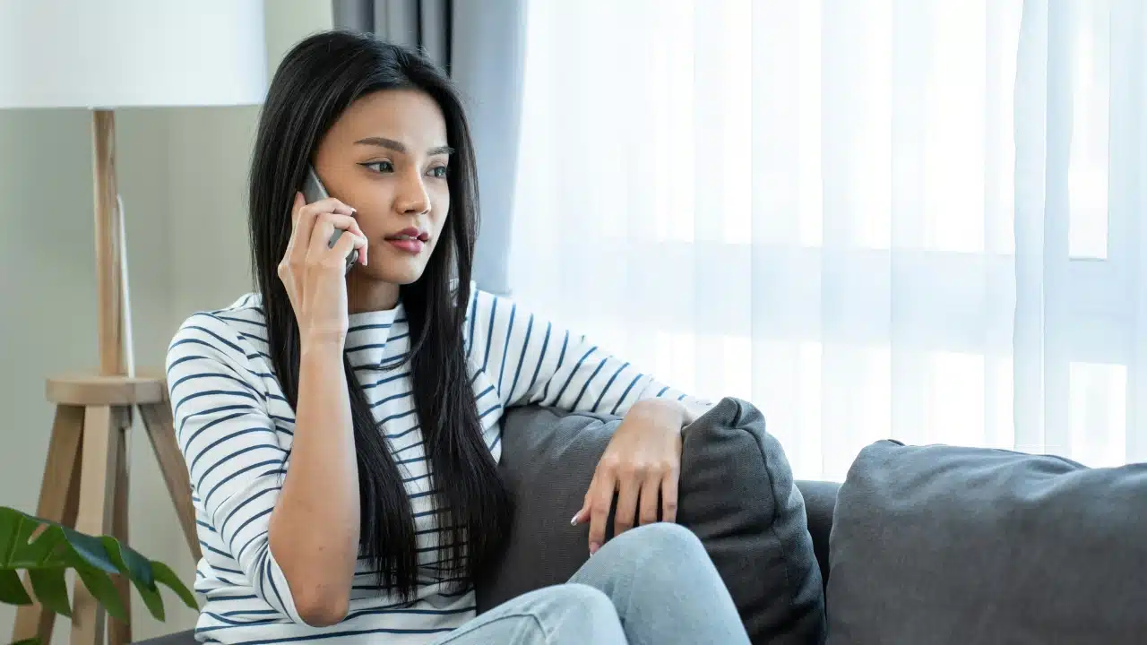 A woman in a striped shirt sits on a gray sofa while holding a smartphone to her ear.