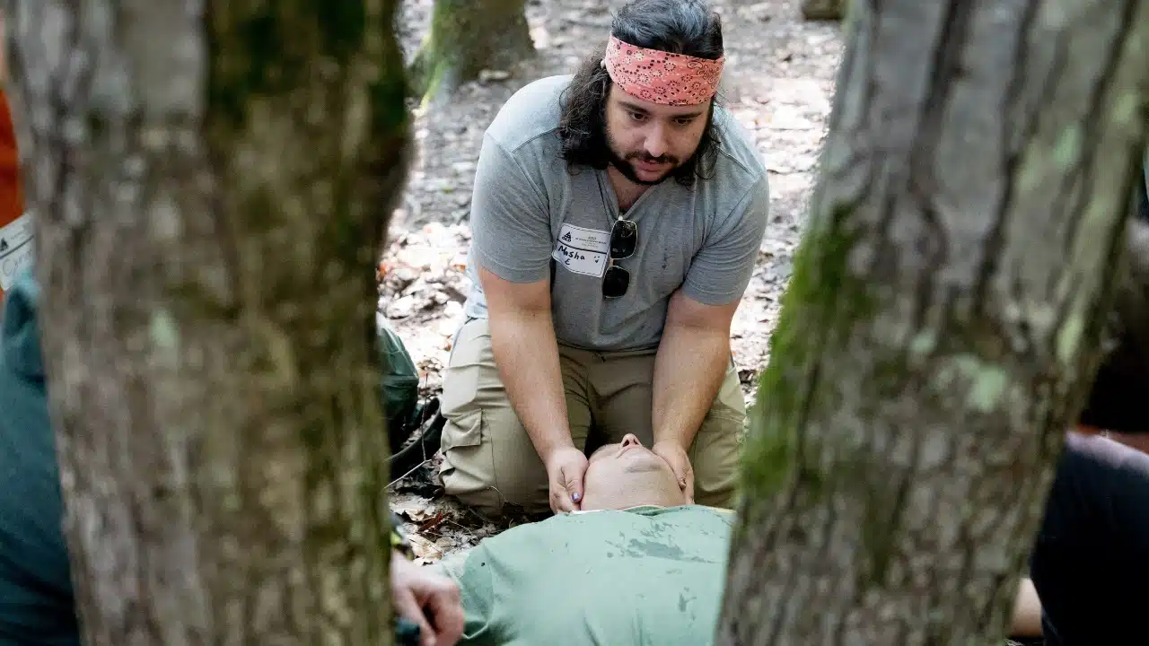 A man in a bandana kneels over another person lying on the forest ground.