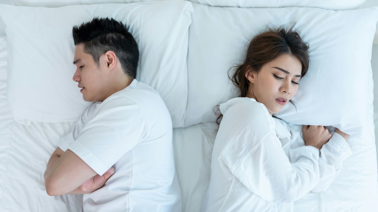 A man and a woman lie in bed facing away from each other with eyes closed.