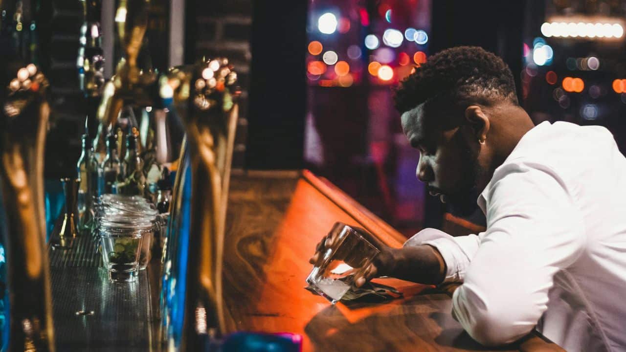 A man in a white shirt leans on a wooden bar counter holding a glass.