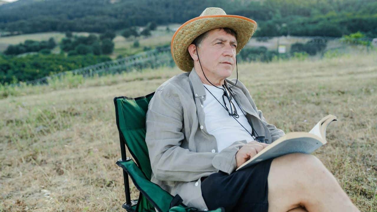 A man wearing a straw hat sits in a folding chair outdoors and holds a book.