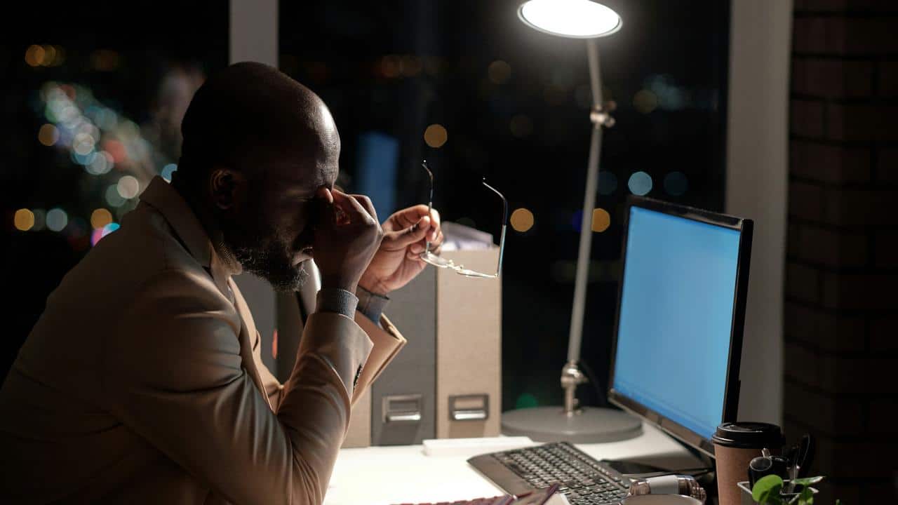 A fatigued man rubbing his eyes while sitting at a desk with a computer at night.