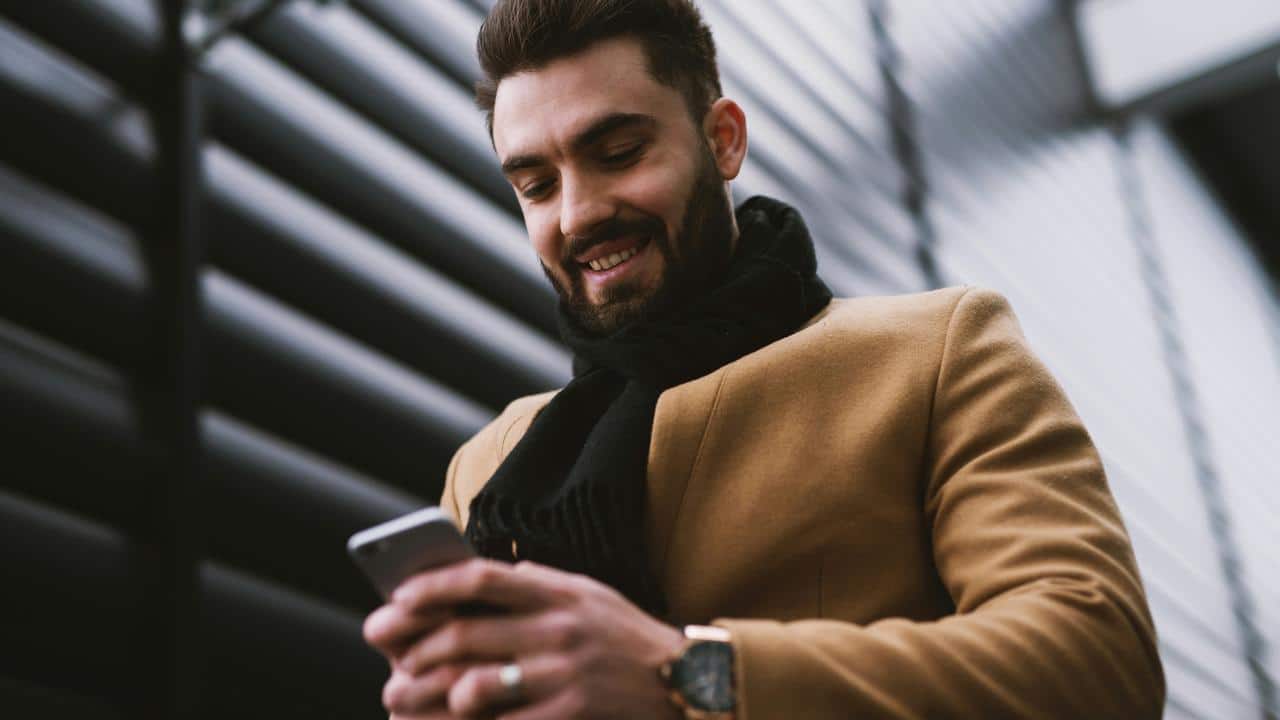 A man wearing a brown coat smiling while texting outdoors.