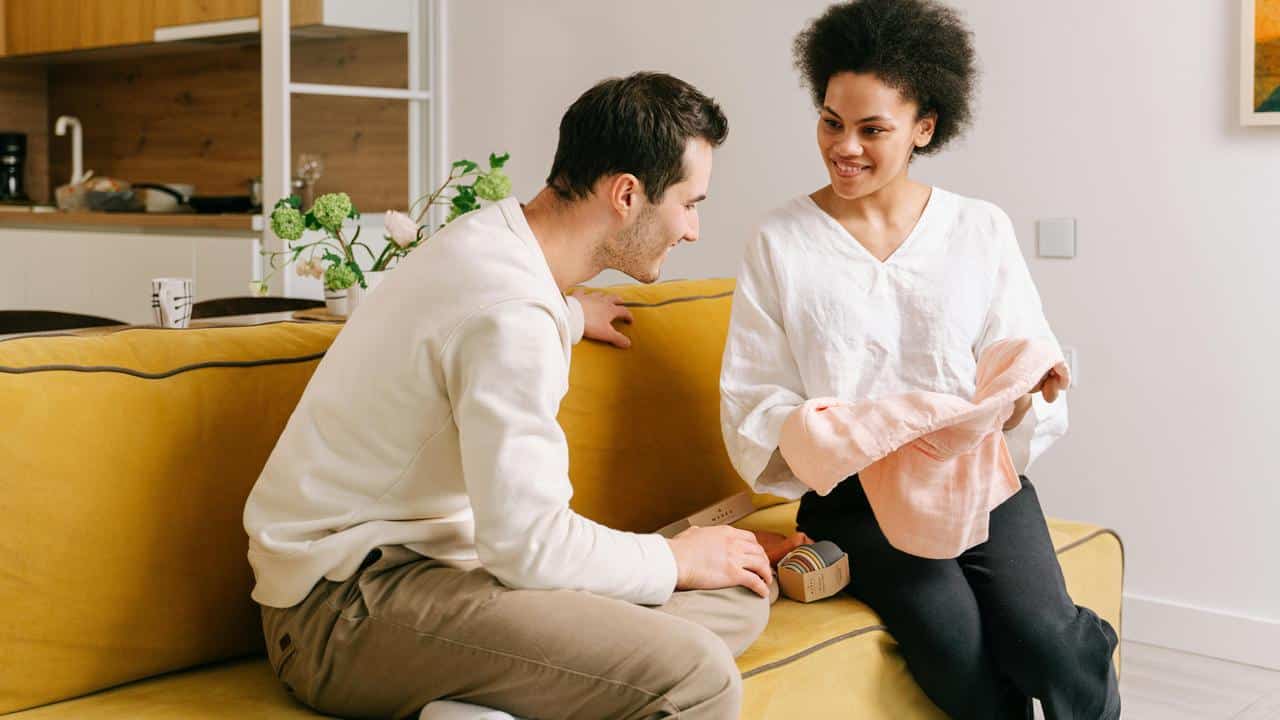 A man and a woman sitting on a couch looking at a piece of clothing together.