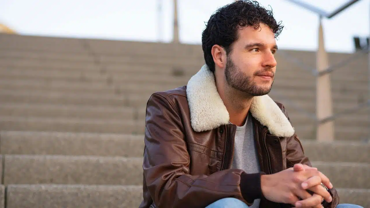 A man sitting on outdoor steps wearing a brown jacket with a shearling collar.