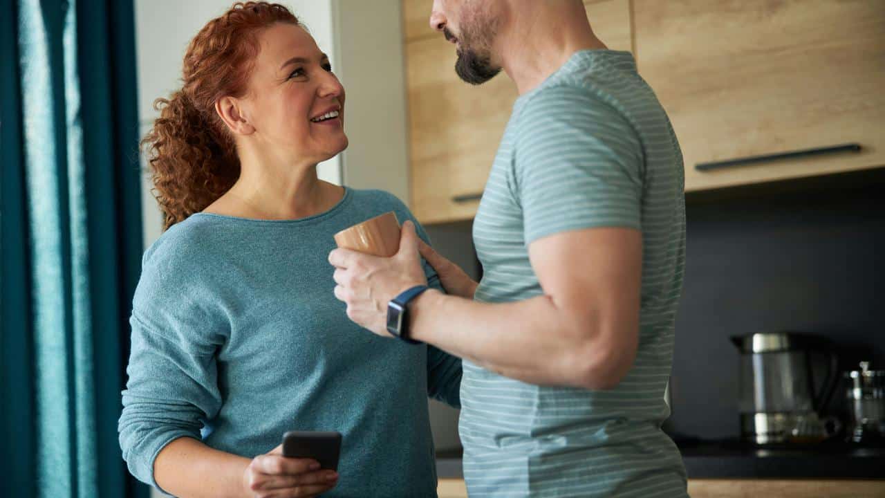 A couple smiles and talks together in a cozy kitchen while holding coffee mugs.