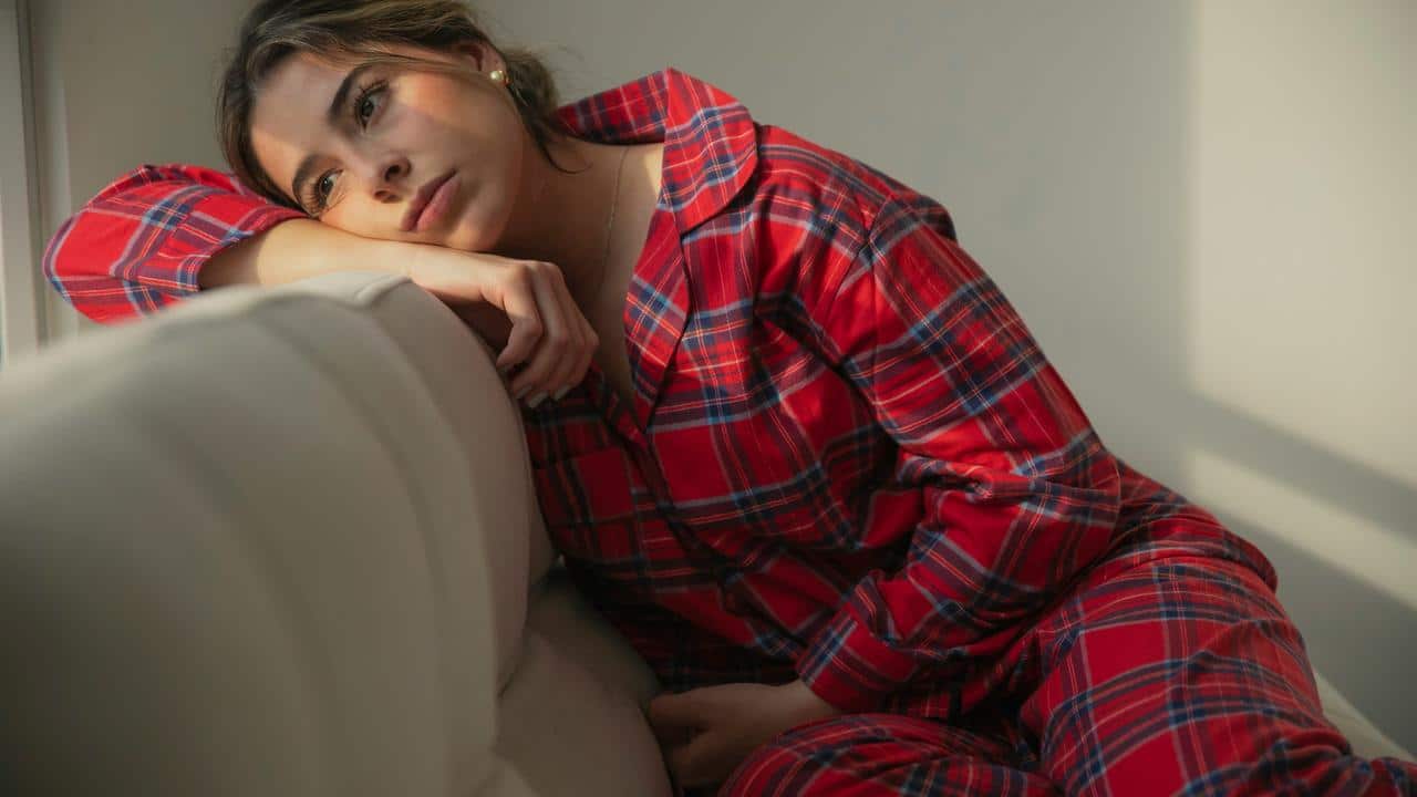 A woman in red plaid pajamas resting her head on a couch and gazing thoughtfully.