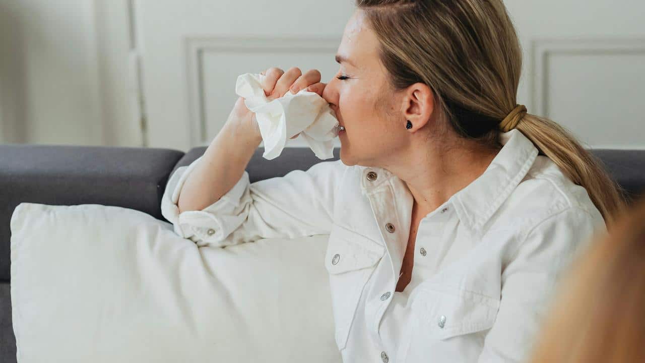 A woman sitting on a couch blowing her nose with a tissue.