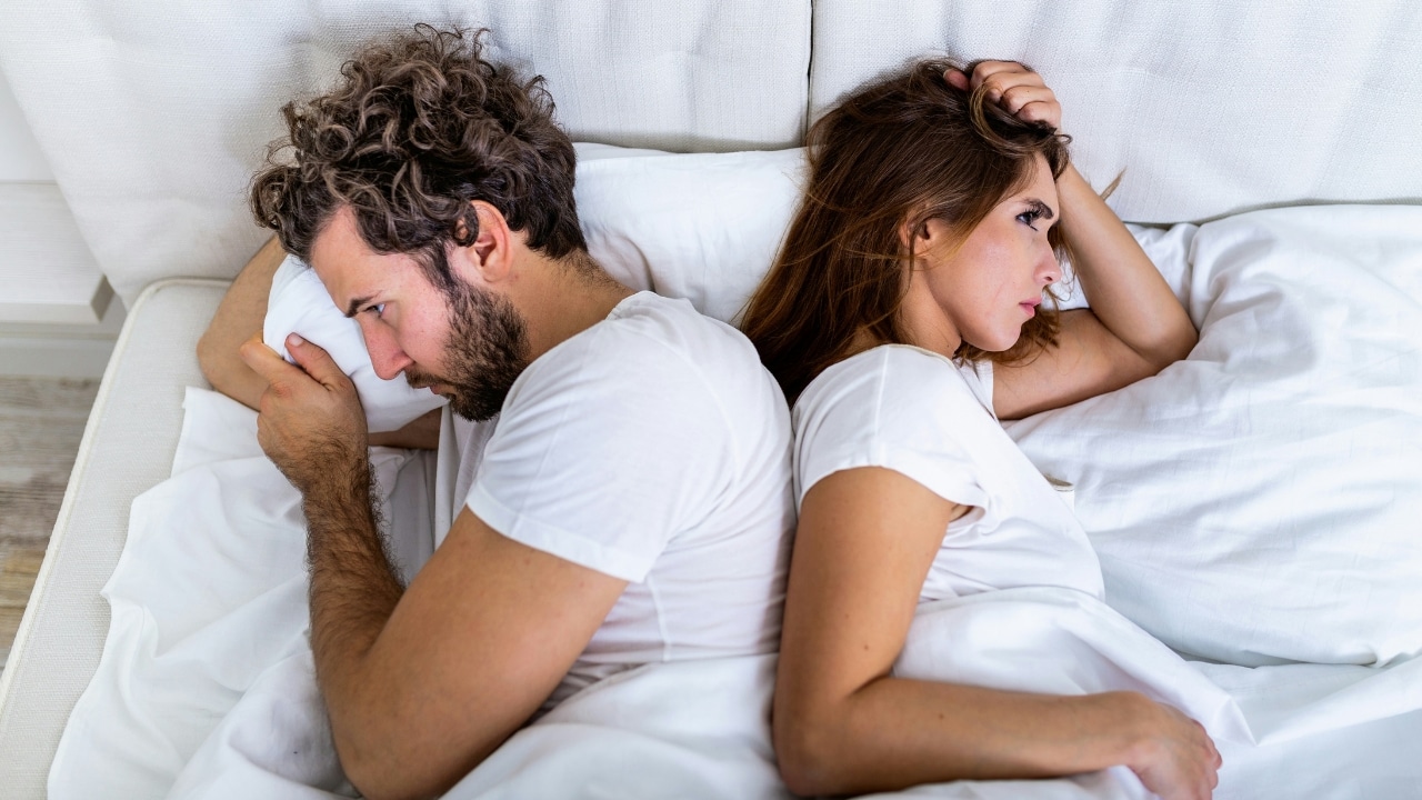 A man and a woman lie in bed back-to-back facing away from each other.