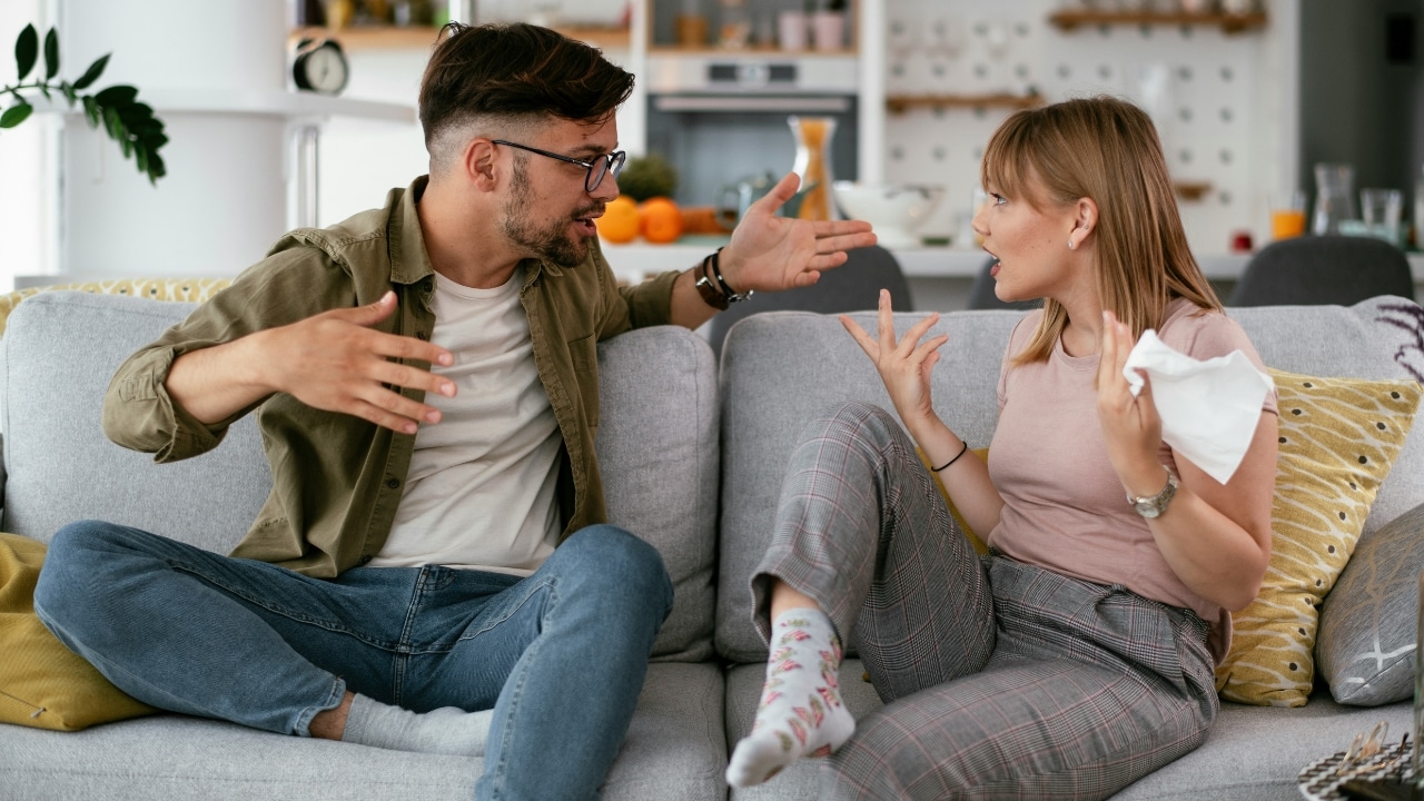 A man and woman sit on a gray sofa, gesturing animatedly toward one another during conversation.