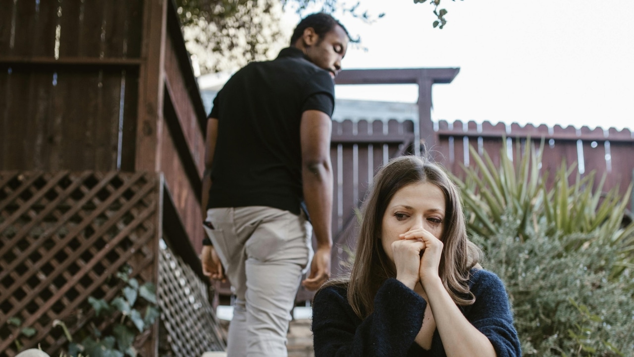 A woman sits outside looking distressed while a man walks away in the background.