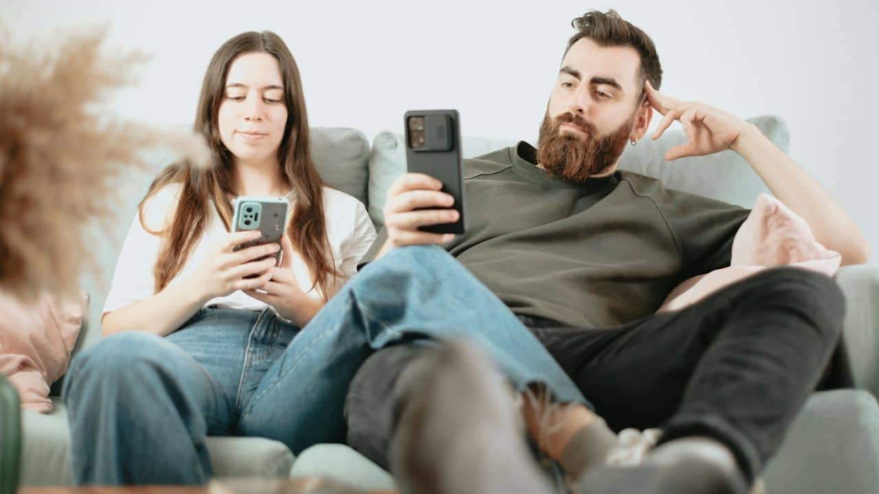 A man and woman sit together on a sofa while both look down at their smartphones.