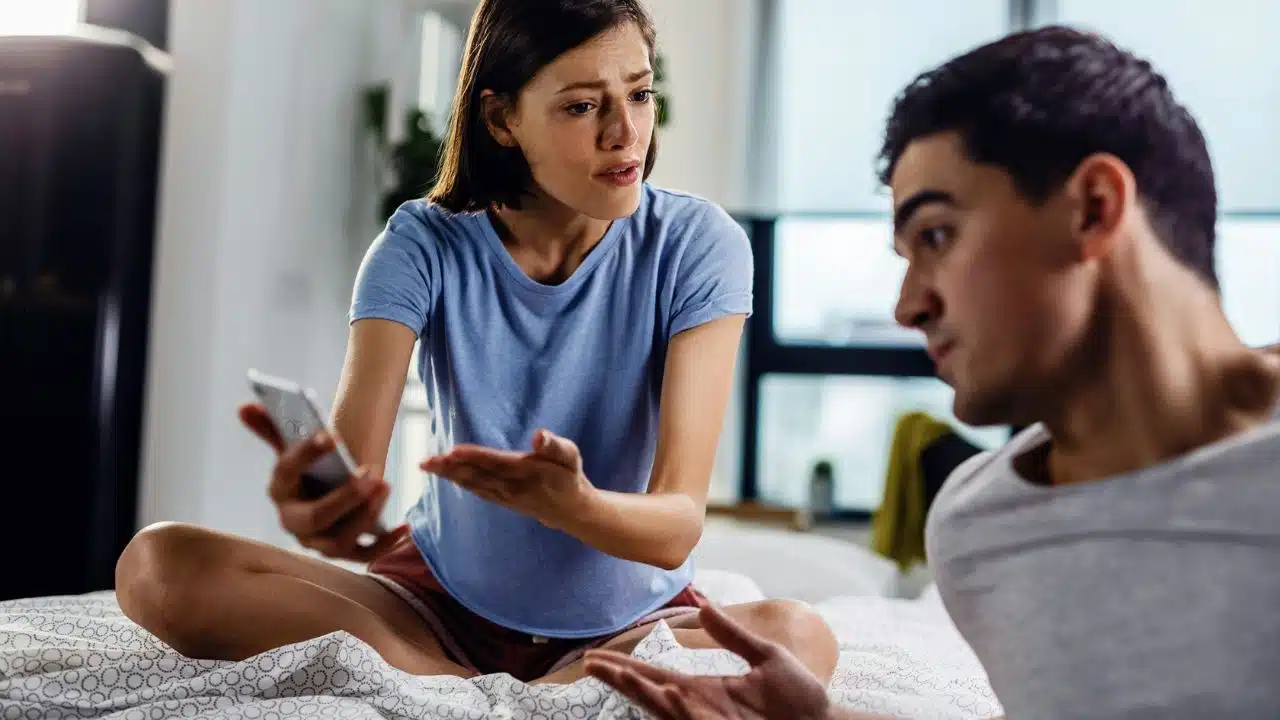 A woman sitting on a bed holds a smartphone and looks at a man gesturing.