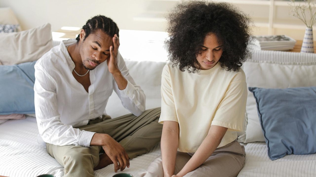 A man with his hand on his head and a woman look down on a sofa.