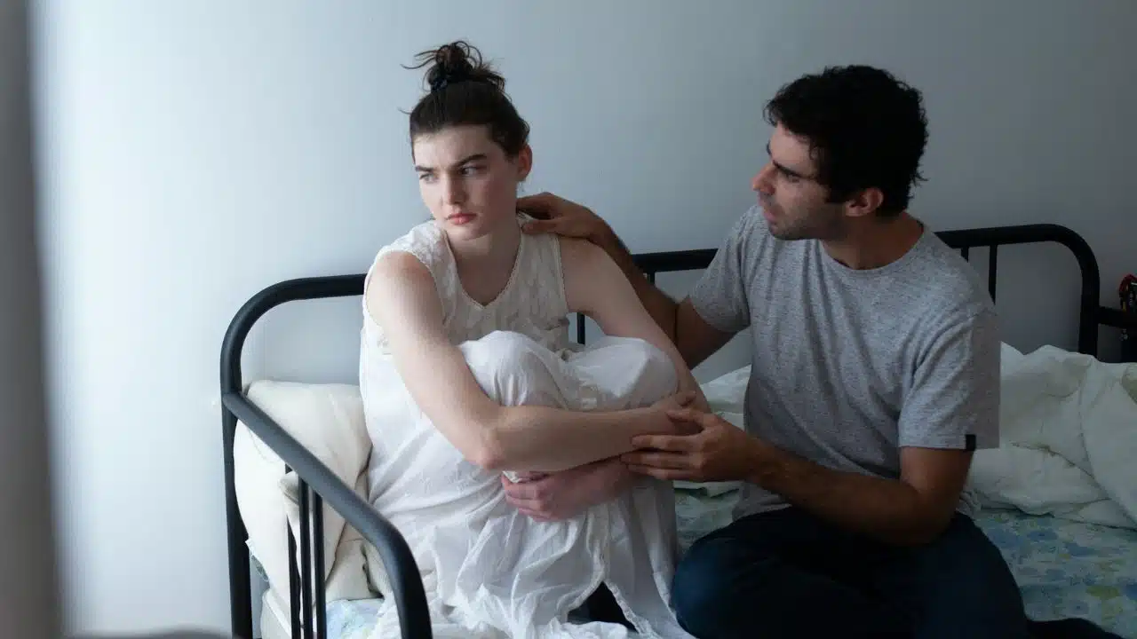 A man reaches out to touch the shoulder of a woman sitting huddled on a bed.