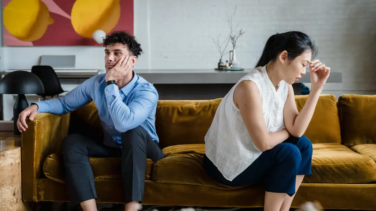 A man and woman sit on a yellow sofa facing away from each other indoors.