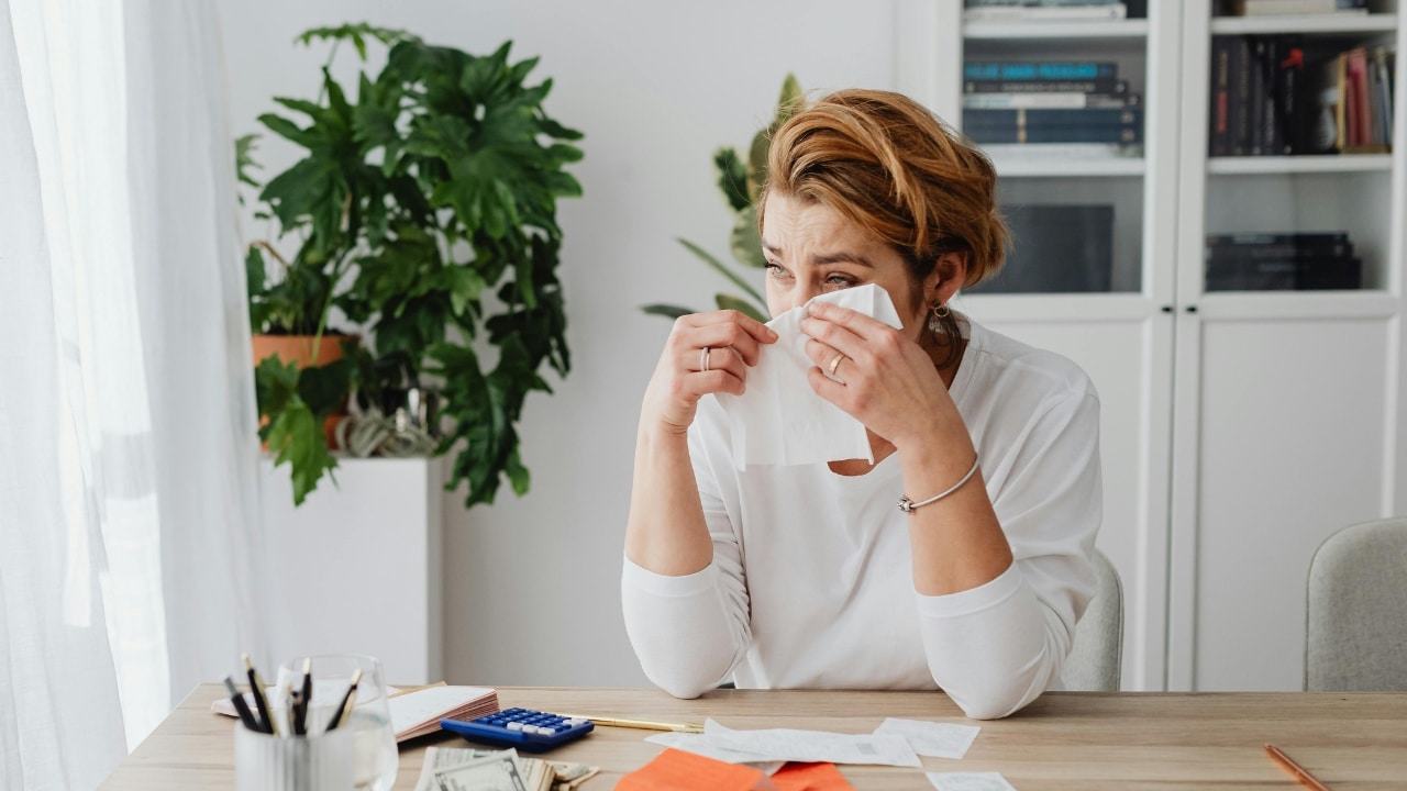 A woman at a desk wipes her nose with a tissue near a calculator and money.