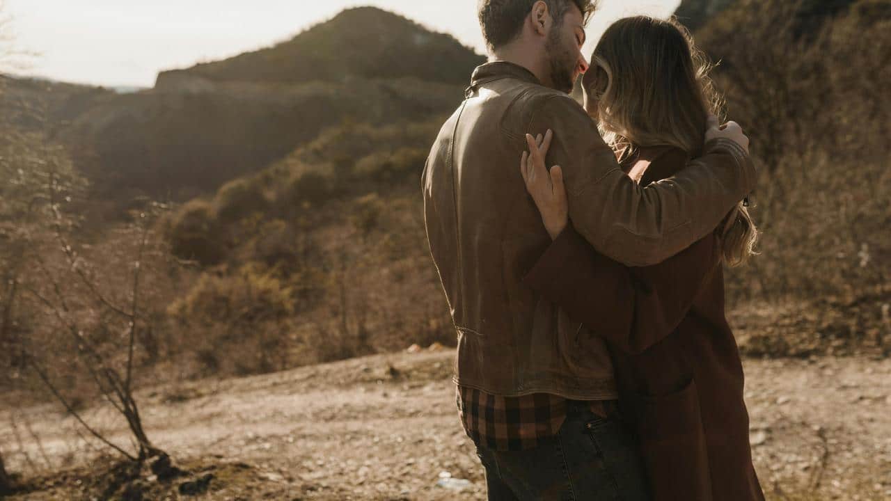 A couple embracing outdoors in a sunlit, mountainous area.