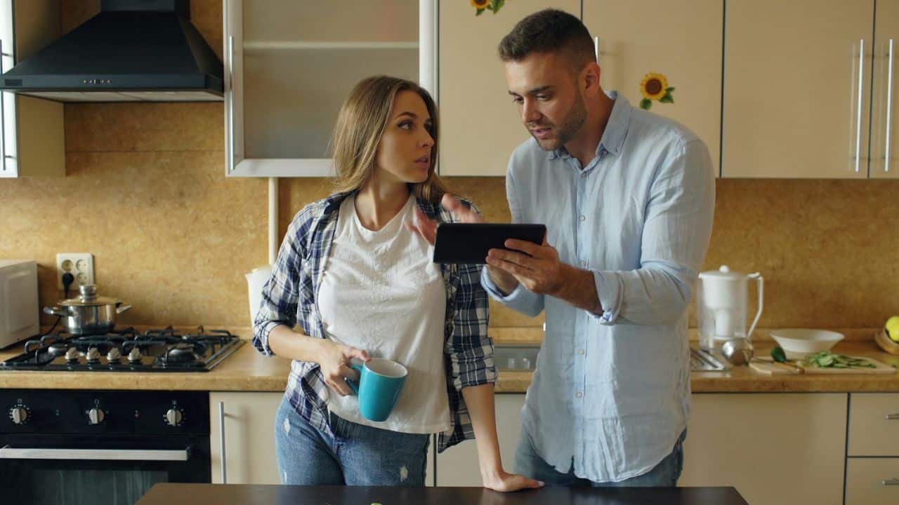 A man and woman standing in a kitchen looking at a tablet while holding a mug.