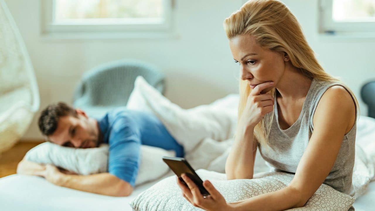 A woman looks at her phone while a man lies in bed in the blurred background.