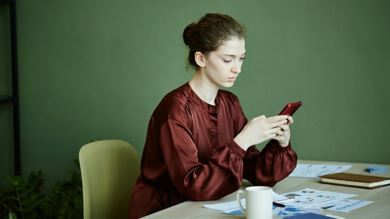A woman sits at a desk with papers and a mug while looking at her phone.