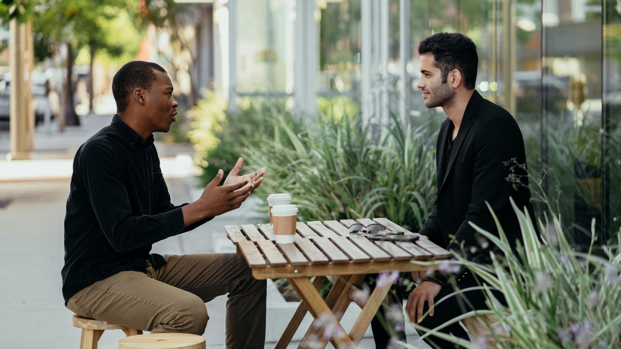 Two men sit at an outdoor wooden table with coffee cups while talking.