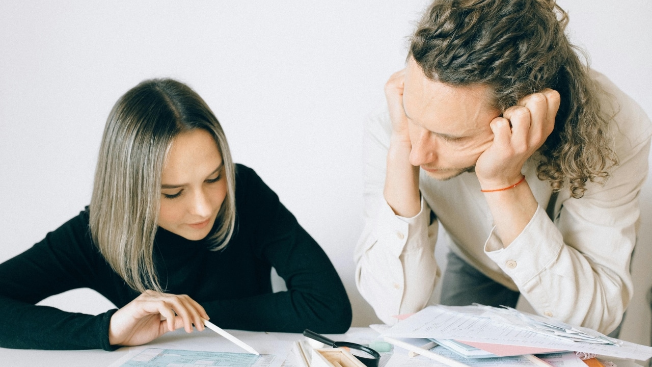 A woman points at a document on a table while a man rests his head.