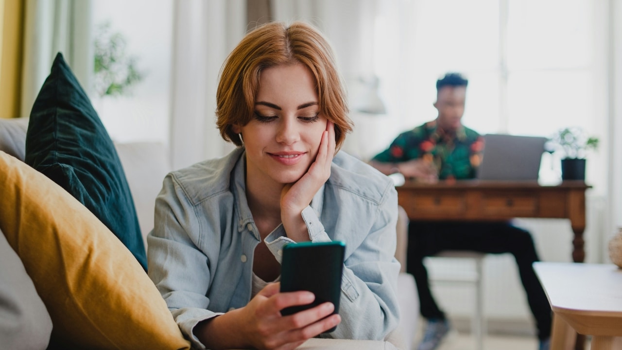A woman smiles while looking at a phone while a man works in background.