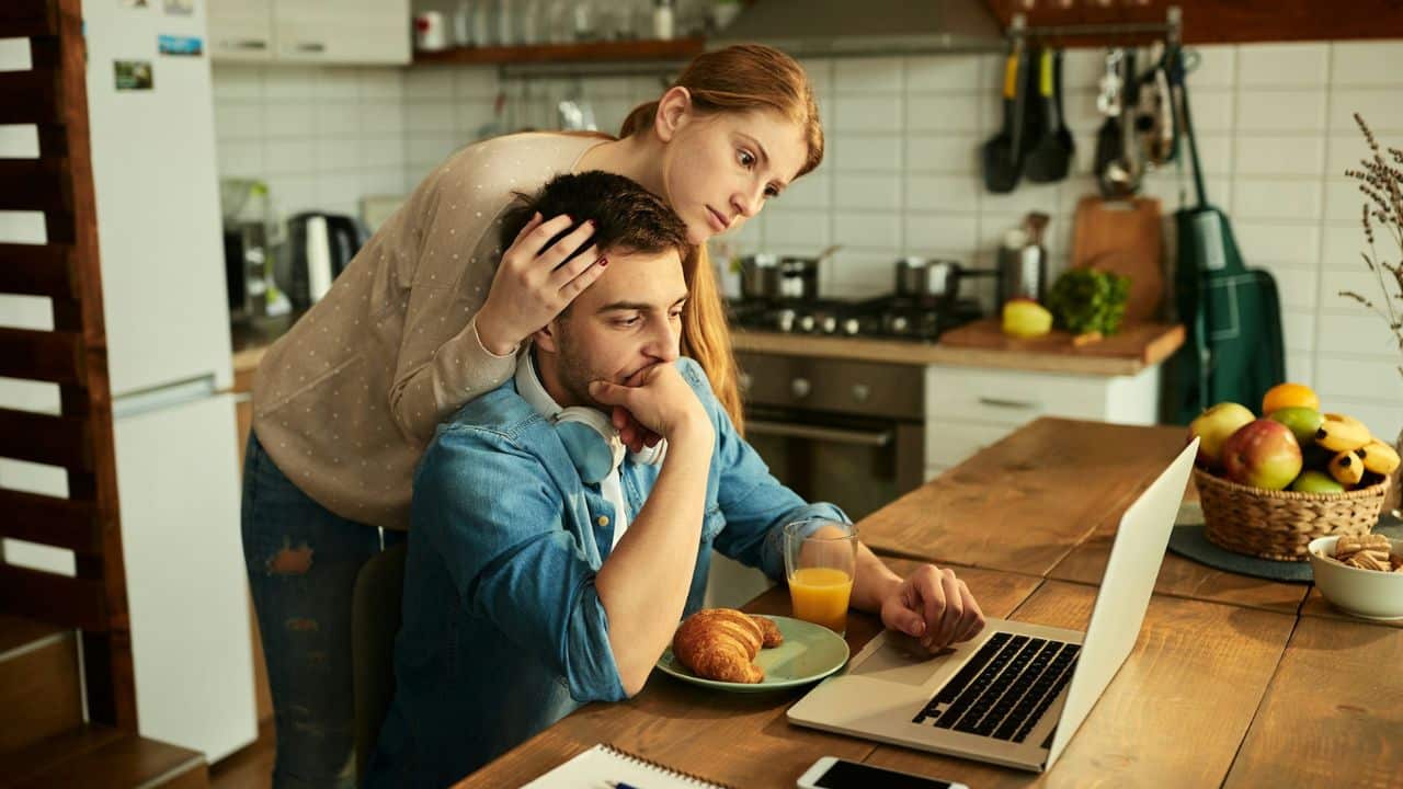A woman stands behind a seated man, resting her hand on his head as they look at a laptop.