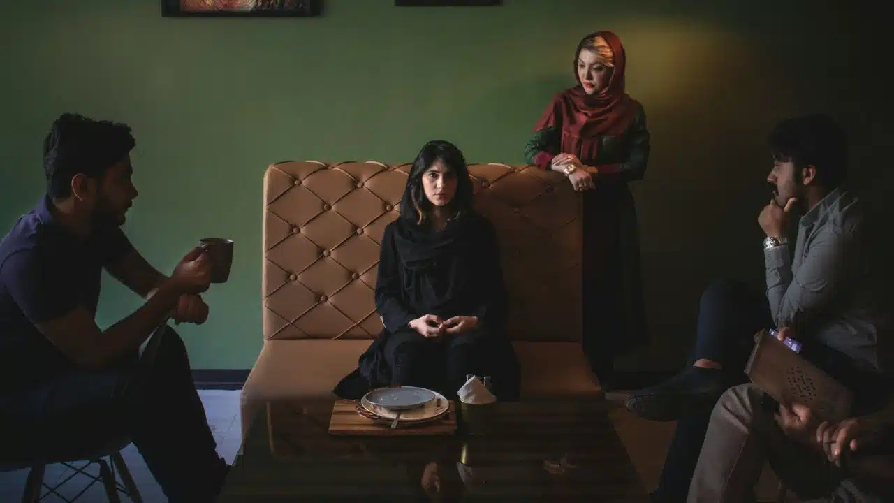 A woman sits on a tufted bench while others stand and sit around her in shadows.