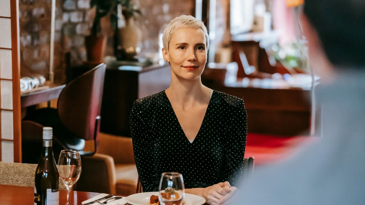 A woman with blonde hair sits at a dinner table looking toward an out-of-focus person.