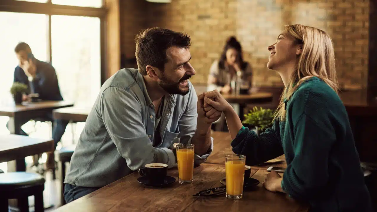 A man and woman sit at a cafe table, smiling and holding each other's hands.
