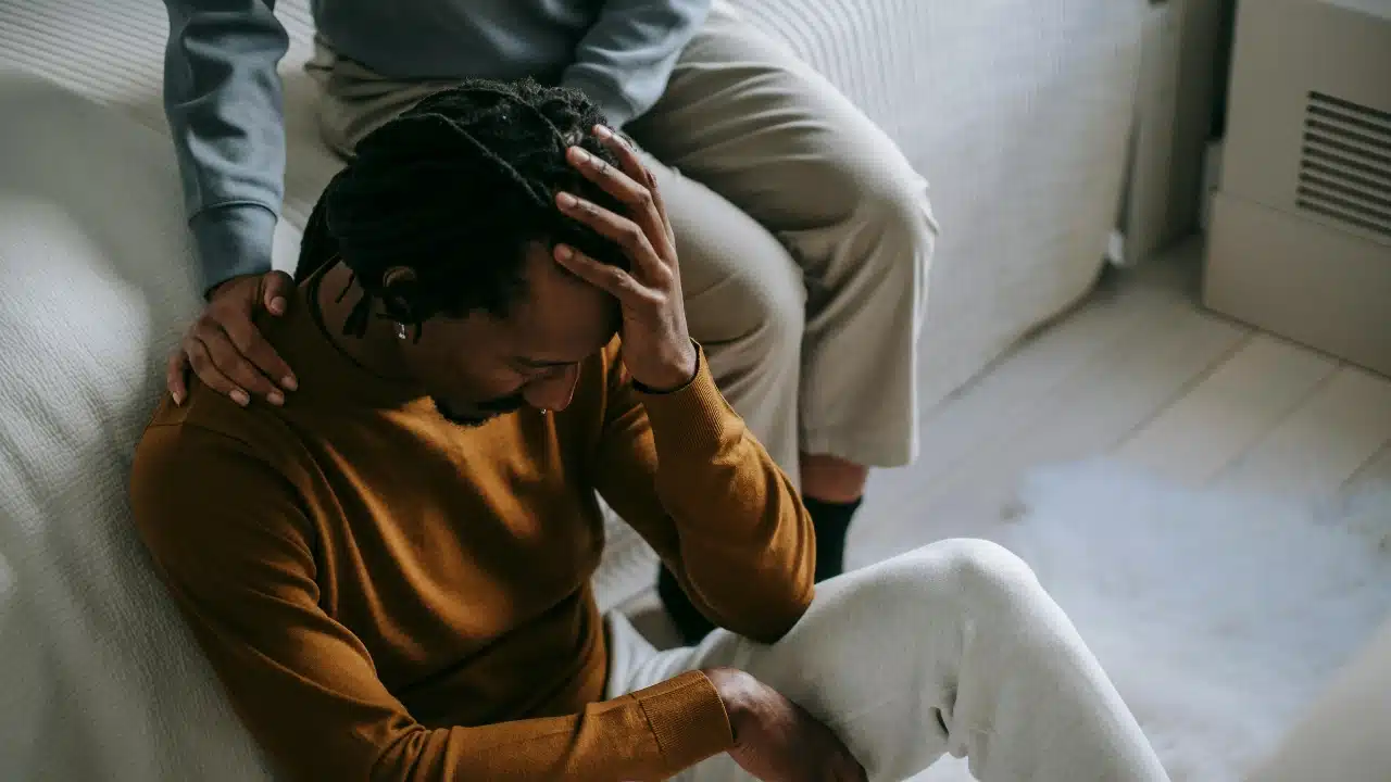 A person places a hand on the shoulder of a man sitting on the floor.