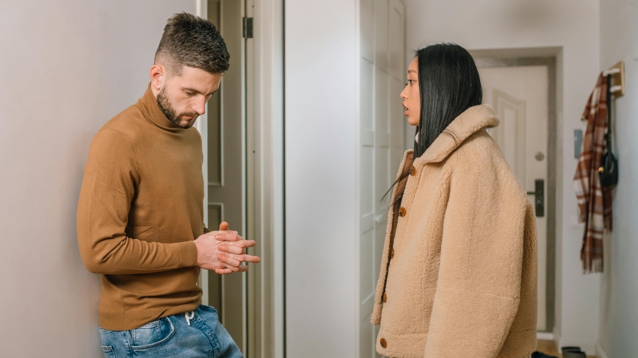 A man in a brown turtleneck looks down while standing near a woman indoors.