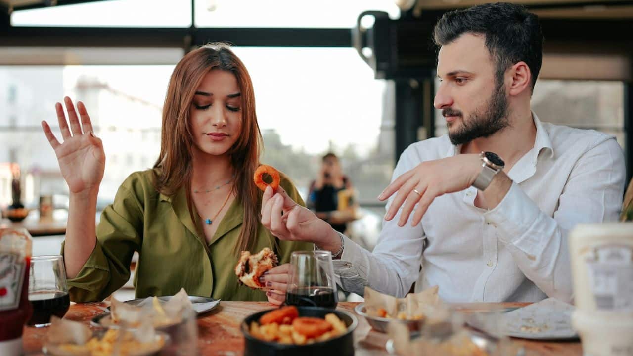 A man holds an onion ring toward a woman who is looking down and waving.